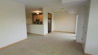 This single-floor space features an open-concept design that seamlessly connects the kitchen and living area, creating a sense of spaciousness and ease of movement. The living area is finished with wall-to-wall neutral-toned carpeting, contributing to a clean and understated aesthetic that suits a variety of décor styles.

The kitchen is partially enclosed by a structural half-wall, which also serves as a breakfast bar or pass-through counter, maintaining visual connectivity with the main living space. Light wood cabinetry lines the kitchen walls, providing ample storage, while stainless steel appliances — including a refrigerator and microwave — add a modern touch. The kitchen is illuminated by a flush-mount ceiling fixture, ensuring functional task lighting.

The living area benefits from an open ceiling with recessed lighting, enhancing the overall brightness of the space. Neutral cream-colored walls run throughout, complementing the light carpeting and creating a cohesive, airy atmosphere. A hallway visible from the main living area suggests access to additional rooms, further adding to the functional layout of this well-organized space.