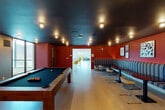 This thoughtfully designed single-floor amenity space offers a rich variety of zones for socializing, dining, and recreation. The game room sets a bold tone with deep red walls, a dark ceiling with industrial-style flush-mount lighting, and light wood flooring. A full-size billiard table anchors the room, complemented by a long striped sectional banquette, lightweight chairs, and a gaming station with dual flat-screen monitors. The adjacent lounge area opens up dramatically, featuring floor-to-ceiling windows framed in teal that flood the space with natural light and reveal sweeping views of the city skyline and waterfront. Built-in upholstered window benches paired with Tulip-style tables and mustard-yellow accent chairs create a relaxed yet sophisticated setting. The dining and kitchen area is a standout, centered around a generous communal table beneath a dramatic cascading chandelier of globe bulbs. A large kitchen island with a dark countertop and teal cabinetry provides ample workspace, while warm wood-paneled walls and professional-grade appliances complete the culinary environment. Throughout, light hardwood floors, curated artwork, and a cohesive contemporary palette tie each distinct zone together into a vibrant and functional shared space.