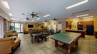 This single-story clubhouse is designed to serve as a versatile community gathering space, thoughtfully laid out to accommodate a variety of social activities. The open main area features large-format tile flooring throughout, crown molding along the ceiling, and multiple ceiling fans that contribute to a comfortable, well-ventilated atmosphere. Recessed and flush-mounted fluorescent lighting keeps the space bright, while rows of wide windows with wood blinds along one wall allow natural light to filter in and provide views of the outdoor surroundings.

The main gathering area is furnished with multiple dark-wood dining tables and chairs, suitable for group meals or events. A ping-pong table occupies a prominent central position, adding a recreational focal point. A dedicated lounge area near the entrance features warm golden leather sofas and accent chairs arranged around a flat-screen television, with decorative brick column accents lending a casual, inviting character to the space.

The fully equipped kitchen features rich wood cabinetry, light granite-style countertops, a center island, pendant lighting, and black appliances including a refrigerator, range, dishwasher, and microwave — well-suited for hosting community events and catered gatherings.