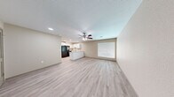 This single-floor residence offers a clean, modern aesthetic defined by light-toned luxury vinyl plank flooring and warm neutral walls that flow consistently throughout the entire space. The open-concept living and dining area is generously proportioned, bathed in natural light through large windows fitted with white horizontal blinds. A ceiling fan with integrated lighting anchors the living space, enhancing both comfort and airflow. The kitchen is thoughtfully designed with crisp white shaker-style cabinetry, light granite countertops, a stainless steel sink, and black appliances including a refrigerator, range, and dishwasher. A peninsula counter creates a natural divide between the kitchen and living area while providing additional workspace. The bedrooms are well-sized, each featuring natural light from windows with blinds and ceiling fans in select rooms. One bedroom includes a louvered double-door closet alongside a six-panel door for added storage. The bathroom is elegantly finished with white cabinetry, granite countertops, a full tub-shower combination clad in large-format gray tile with a built-in niche, and light wood-look flooring. Recessed lighting and a textured ceiling complete the refined, move-in-ready interior throughout.