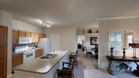 This single-floor residence in Lewisville, Texas offers a well-designed open-concept layout that seamlessly connects the kitchen, dining, and living areas. The kitchen is equipped with oak cabinetry, a white refrigerator, a white range, and a tile backsplash. A generous peninsula counter with a double-basin sink extends into the space, providing ample bar-style seating — ideal for casual dining or entertaining. Track lighting illuminates the kitchen area, ensuring bright and functional workspace. Moving toward the living and dining areas, the flooring transitions to plush carpet, adding warmth and comfort underfoot. Crown molding lines the ceilings throughout, lending a refined architectural detail to the interior. The dining area features a round table with cushioned chairs and is accented by a built-in shelving unit, perfect for displaying décor. A hallway visible from the main living space leads to additional rooms, offering a sense of depth and privacy. The neutral color palette of warm beige tones throughout creates a cohesive and inviting ambiance, making this Lewisville property a comfortable and practical single-level home.