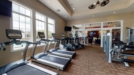 Located in Westerville, Ohio, this single-floor fitness center offers a comprehensive workout facility designed to support a wide range of exercise routines. The space is bathed in natural light, courtesy of large, multi-pane windows along multiple walls that provide open views of the surrounding community. Elevated ceilings with a tray detail and ceiling fans contribute to an airy, spacious atmosphere throughout.

The cardio zone lines the windowed perimeter and features a row of treadmills, elliptical trainers branded by TRUE Fitness, and stationary bikes, each positioned for natural light and accompanied by wall-mounted flat-screen televisions. Adjacent to the cardio area, a strength training section houses a selection of cable and resistance machines suited for a full-body workout.

A dedicated free weights area includes a tiered dumbbell rack, a workout bench, medicine balls, and stability balls, all set on protective rubber flooring with full-length mirrors to assist in proper form. An entryway utility nook provides dark wood cabinetry with granite countertops, a water dispenser, paper towel stations, and wall-mounted suspension training straps and accessories. The warm, neutral color palette, carpeted cardio zones, and recessed lighting create a clean, welcoming environment for residents seeking an on-site fitness experience.