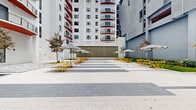This open-air courtyard in Miami, FL offers a dynamic urban gathering space defined by bold design choices and thoughtful landscaping. The ground plane features a geometric pattern of alternating light and dark stone pavers that stretch across the expansive plaza, creating a strong visual rhythm underfoot. Bright yellow folding chairs and tables are arranged throughout the courtyard, shaded by classic black-and-white striped umbrellas that add a playful, café-like character to the space.

Lush strips of artificial turf border the plaza, complemented by manicured garden beds and young trees that introduce natural elements into the urban setting. A standout architectural feature is a striking accent wall clad floor-to-ceiling in bold black-and-white patterned tiles, incorporating a sleek water feature at its base that adds both visual interest and a soothing ambiance.

The courtyard is framed on multiple sides by modern multi-story buildings with white facades, red accents, and balconies that overlook the space below. A covered parking entry with a contemporary louvered canopy anchors one edge of the courtyard. Decorative steel bollards guide pedestrian flow, and the entire space is open to the South Florida sky, bathing the area in abundant natural light and creating a welcoming environment for gathering, dining, and relaxation.