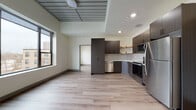 Located at 410 W Broadway St in Mount Pleasant, Michigan, this thoughtfully designed unit offers a contemporary living experience across a single floor. The open-concept living and kitchen area is anchored by light-toned hardwood-style flooring that flows seamlessly throughout the space, creating a cohesive and airy feel.

The kitchen is a standout feature, outfitted with dark, textured cabinetry, open floating shelves, a full suite of stainless steel appliances including a refrigerator, range, built-in microwave, and dishwasher, as well as a clean white tile backsplash and light countertops. The kitchen transitions naturally into the living area, which benefits from elevated ceilings with a distinctive panel detail and recessed lighting that adds to the modern aesthetic.

Large, dark-framed windows in the living area and bedroom flood the space with natural light and offer open views of the surrounding landscape and waterway below. The bedroom is a calm, well-proportioned retreat with its own generous window and roller shade. The bathroom features a double-sink vanity with an illuminated mirror, dark wood cabinetry, white quartz countertops, and a full soaking tub with shower. Every detail reflects a clean, modern design sensibility.