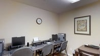 This single-floor space functions as a dedicated business center, offering a focused environment for productivity and administrative tasks. The room features neutral beige walls paired with a textured popcorn ceiling, creating a modest and utilitarian ambiance. Overhead lighting provides adequate illumination throughout the space.

A long, dark-surfaced workstation desk runs along one wall, accommodating multiple workstations. The setup includes desktop computers with monitors, keyboards, and a shared printer, making the space well-suited for tasks such as document printing, browsing, and general computer use. Several cushioned chairs with metal frames are positioned along the desk, offering seating for multiple users simultaneously.

A framed wall clock is mounted on the wall, providing easy time reference for occupants. On the adjacent wall, a framed decorative piece adds a subtle touch of visual interest. A small side table or cabinet is partially visible near the entrance, contributing additional utility to the room.

The overall design is straightforward and functional, prioritizing practicality over aesthetics. The space is well-suited for use as a shared business or computer center, such as those commonly found in hospitality or communal facilities.