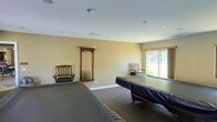This single-floor space is centered around a spacious billiards room designed for recreational enjoyment. The room is generously sized, comfortably accommodating multiple pool tables with room to maneuver around each one. The walls are finished in a warm, neutral tone that contributes to a relaxed and welcoming atmosphere throughout.

Recessed lighting in the white ceiling provides even, functional illumination across the playing surfaces, complemented by an abundance of natural light streaming in through a large sliding glass door. The door offers pleasant views of the surrounding greenery outdoors, enhancing the overall ambiance of the room.

A wall-mounted wooden cue rack keeps billiard cues neatly organized and readily accessible, serving as both a practical storage solution and a tasteful decorative element. Two wooden chairs are positioned along the walls, offering comfortable seating for players waiting their turn. The floor is finished with wall-to-wall carpet in a neutral tone, adding warmth underfoot and reducing noise within the space.

Overall, this billiards room strikes a balance between functionality and comfort, making it a well-appointed destination for leisure and entertainment.