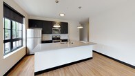 Unit 217 is a thoughtfully designed single-floor residence that blends contemporary aesthetics with functional living. Light wood flooring runs consistently throughout the space, complemented by crisp white walls and bold black baseboards that create a clean, modern contrast.

The open-concept living and dining area is generously proportioned, bathed in natural light through large black-framed grid windows that frame views of the surrounding neighborhood. A glass sliding door provides additional access to the outdoors, further enhancing the sense of openness.

The kitchen is a standout feature, anchored by a spacious waterfall island with quartz countertops and an integrated sink. Dark cabinetry reaches toward the ceiling, paired with a classic white subway tile backsplash and stainless steel appliances including a refrigerator, range, and microwave. Industrial-style pendant lights add a stylish finishing touch.

The bedrooms are well-proportioned, each featuring the same warm wood flooring and black-framed windows that invite ample natural light and outdoor views.

Two bathrooms serve the space. The primary bathroom features floor-to-ceiling dark gray tile, a glass-enclosed tub and shower combination, and an illuminated mirror. The secondary bathroom offers a clean, minimal design with a soaking tub and shower, a gray vanity with dark countertop, and a lighted mirror. Both bathrooms continue the warm wood flooring found throughout.