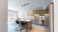 Located at 2050 W Ogden Ave in Chicago's West Side, this thoughtfully designed one-bedroom unit combines contemporary finishes with industrial-inspired architectural elements. The exposed concrete ceiling and light wood-plank flooring run throughout, creating a cohesive, modern aesthetic.

The open-concept kitchen is a standout feature, outfitted with warm wood-tone cabinetry, under-cabinet LED lighting, white quartz countertops, a tile backsplash, and a full suite of stainless steel appliances including a range, microwave, dishwasher, and refrigerator. A central kitchen island with bar seating connects seamlessly to the living area, which is bathed in natural light from floor-to-ceiling windows. These windows open to a private balcony offering elevated views of the surrounding Chicago neighborhood.

The bedroom features the same exposed concrete ceiling with a structural column detail, built-in closet storage with clean white panel doors, and ample natural light from a large window. The bathroom is finished with marble-look tile flooring, a wall-mounted sink, an illuminated mirror, and crisp white walls, delivering a clean and functional design. The overall ambiance of the space is bright, urban, and refined.