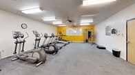 This fitness center at 739 S 300 W in Salt Lake City, Utah, offers a dedicated single-floor exercise room designed to accommodate a comprehensive range of workout activities. The space is outfitted with multiple treadmills and elliptical machines arranged in a row, providing ample cardio training options. A cable strength machine, a power tower for bodyweight exercises, a stationary bike, and a free weight area complete with dumbbells and exercise balls round out the strength and cross-training capabilities. A full-length mirrored wall enhances the sense of openness and allows users to monitor their form during workouts. The room features neutral white walls accented by a bold yellow feature wall, adding a vibrant, energizing atmosphere. Gray carpet flooring runs throughout, and the textured ceiling is equipped with multiple fluorescent light fixtures, ensuring bright, consistent illumination. Ceiling fans provide additional air circulation throughout the space. A window along the yellow accent wall allows for natural light. The room also includes a separate alcove area with additional equipment, offering a more private workout zone. The overall layout is functional and organized, providing a comfortable environment for residents to pursue their fitness goals.