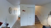 This two-story residence offers a straightforward and functional layout spread across two levels, connected by a carpeted staircase. The interior is finished in a clean, neutral palette, with white walls and light-toned ceilings that contribute to a bright and airy atmosphere throughout.

The main level showcases wall-to-wall carpeting in the common areas, providing a warm and comfortable underfoot feel. A closet with a paneled white door serves as practical built-in storage near the staircase. Decorative touches, including circular wall art and a distinctive red key ornament, add subtle character to the space. A glimpse into an adjoining area reveals hardwood-style flooring, natural light filtering through a window, and additional furnishings, suggesting a dedicated living or dining zone.

The staircase, finished with carpet treads and a painted baseboard, leads to the upper floor, where additional living spaces are accessible. The textured ceiling finish is consistent across both levels, reinforcing a cohesive design aesthetic. Overall, the space presents a well-maintained, move-in-ready environment with a neutral, versatile interior that lends itself easily to personalization.