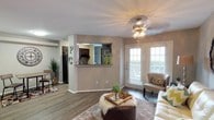 This single-floor residence offers a thoughtfully designed open-concept layout that seamlessly connects the living and kitchen areas. The living room is a welcoming focal point, featuring warm hardwood-style flooring, neutral-toned walls, and a decorative chandelier that adds a touch of elegance overhead. Natural light floods the space through large windows fitted with white shutters and a set of French doors, creating a bright and airy atmosphere. A pass-through window cut into the dividing wall connects the living area to the kitchen, promoting an open feel while maintaining a defined separation between spaces. The kitchen is visible through this pass-through and features dark cabinetry, a range, and a refrigerator, offering a functional and well-equipped cooking space. The overall ambiance is warm and inviting, enhanced by thoughtful décor accents, a plush area rug, and a mix of casual and refined furnishings. The layout is efficient and practical, with clear sightlines throughout the main living area. Additional rooms are accessible from the main space, further expanding the functional footprint of this single-story residence.