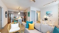 This single-floor residence showcases a thoughtfully designed open-concept layout that seamlessly connects the kitchen, dining, and living areas, creating a bright and cohesive living environment. The kitchen is a functional centerpiece, featuring sleek gray flat-front cabinetry extending to the ceiling, white quartz countertops, a subway tile backsplash, and a full suite of stainless steel appliances. A kitchen island with pendant lighting serves as both a prep space and a casual dining spot, complemented by vibrant teal metal barstools that add a playful pop of color. Light wood-style flooring runs throughout, enhancing the airy feel of the space. The dining area, positioned adjacent to the kitchen, accommodates a compact table with seating for four beneath track lighting. The living room is anchored by a ceiling fan, multiple windows dressed in botanical-print curtains that allow ample natural light, and a cozy mix of a gray sofa, woven rattan accent chair, and layered area rugs. Coastal-inspired décor, including a bold flamingo wall canvas, reinforces a relaxed, resort-like ambiance throughout the residence.