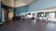 This fitness center in American Fork, Utah, is a thoughtfully designed single-floor facility built to support a comprehensive workout experience. The space is characterized by dramatically high vaulted ceilings that lend an open, airy feel, while a bold teal and white color scheme gives the interior a clean, modern aesthetic. Dark rubber flooring throughout provides a durable, slip-resistant surface well-suited for exercise.

The cardio section features multiple treadmills positioned in front of large windows that allow ample natural light to fill the space and offer views of the surrounding area. A wall-mounted television accompanies this zone for added entertainment during workouts. Additional cardio equipment includes a StairMaster and an elliptical machine, positioned to make efficient use of the floor space.

The strength training area is equally well-appointed, with a full dumbbell rack stocked with a wide range of weights alongside medicine balls and benches. Multiple cable and pulley machines, as well as selectorized strength stations, are distributed throughout the space. Large mirrors mounted along the walls enhance the sense of depth, assist with form, and visually expand the room. The overall layout is open and uncluttered, allowing users to move freely between equipment zones.