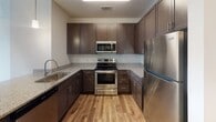 Located at 15708 WC Main St in Midlothian, Virginia, this single-floor unit presents a clean, contemporary design with consistent finishes throughout. The open-concept layout connects the living area to the kitchen, creating a cohesive and functional flow. The kitchen is well-equipped, featuring rich dark-brown shaker-style cabinetry, granite countertops, a stainless steel refrigerator, electric range, and over-the-range microwave. A peninsula with pendant lighting serves as a natural divider between the kitchen and the living space, adding both visual interest and additional prep or seating area.

Hardwood-style flooring runs through the main living areas and kitchen, offering a warm, polished aesthetic that is easy to maintain. The bedrooms include ceiling fans and ample natural light provided by large windows with blinds. One bedroom features hardwood-style flooring, while another is finished with plush carpeting, offering distinct comfort options.

The bathroom maintains the same design language as the kitchen, with dark cabinetry, granite countertops, and hardwood-style tile flooring. A well-lit vanity mirror and open wire shelving near the entrance offer practical storage. Throughout the unit, neutral wall tones and bright recessed lighting contribute to an airy, modern ambiance.