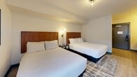 Located at 461 Munson Ave in Traverse City, Michigan, this well-appointed guest room offers a thoughtfully designed layout across a single floor. The bedroom accommodates two queen beds, each featuring warm wood headboards, crisp white bedding, and wall-mounted globe sconce lighting positioned between them for both style and function. Light wood-style flooring runs throughout the room, complemented by a patterned area rug in cool gray and blue tones. A window along one wall allows natural light to filter in, offering views of the surrounding area, while a wall-unit HVAC system ensures year-round climate comfort. A work desk and chair provide a functional workspace within the room. The unit includes two bathrooms, dividing convenience efficiently. The vanity area features a floating wood-trimmed countertop with an integrated sink, a large illuminated mirror, open shelving for toiletries, and a compact mini-refrigerator tucked beneath. The second bathroom is a dedicated wet room with a glass-enclosed shower stall stocked with amenity dispensers, a toilet, and overhead shelving for fresh towels. Light stone-look tile flooring ties both bathroom areas together cohesively. The overall aesthetic is clean, modern, and functional — well-suited for travelers seeking comfortable, practical accommodations in Traverse City.