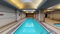 This single-level indoor aquatic facility is thoughtfully designed to accommodate a variety of swimming and leisure activities. The centerpiece is a long, narrow lap pool with crystal-clear turquoise water, bordered by slip-resistant mosaic tile decking that extends throughout the space. Overhead, a recessed skylight runs along the length of the lap pool, flooding the area with natural light and enhancing the open, airy atmosphere complemented by warm recessed and cove lighting.

Adjacent to the lap pool, a circular hot tub offers a dedicated relaxation area, while a separate wading pool provides a shallower option for leisurely use. Structural columns punctuate the space and help define distinct zones within the facility. The walls are finished in a neutral palette of white and grey mosaic tile, accented with decorative multi-tone tile banding that adds visual interest at mid-height.

Poolside lounge chairs and small table-and-chair sets are positioned along the perimeter, offering comfortable spots to rest between swims. Potted plants add touches of greenery near the far end of the lap pool. Multiple doorways lead to changing rooms and additional amenities, while a drinking fountain is conveniently positioned along one wall. The overall ambiance is clean, functional, and well-maintained.