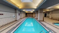 This single-level indoor aquatic facility is thoughtfully designed to accommodate a variety of swimming and leisure activities. The centerpiece is a long, narrow lap pool with crystal-clear turquoise water, bordered by slip-resistant mosaic tile decking that extends throughout the space. Overhead, a recessed skylight runs along the length of the lap pool, flooding the area with natural light and enhancing the open, airy atmosphere complemented by warm recessed and cove lighting.

Adjacent to the lap pool, a circular hot tub offers a dedicated relaxation area, while a separate wading pool provides a shallower option for leisurely use. Structural columns punctuate the space and help define distinct zones within the facility. The walls are finished in a neutral palette of white and grey mosaic tile, accented with decorative multi-tone tile banding that adds visual interest at mid-height.

Poolside lounge chairs and small table-and-chair sets are positioned along the perimeter, offering comfortable spots to rest between swims. Potted plants add touches of greenery near the far end of the lap pool. Multiple doorways lead to changing rooms and additional amenities, while a drinking fountain is conveniently positioned along one wall. The overall ambiance is clean, functional, and well-maintained.