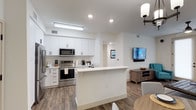 This single-floor space showcases a clean, contemporary design centered around an open-concept layout that connects the kitchen and living area seamlessly. The kitchen is outfitted with stainless steel appliances, including a refrigerator, electric range, and over-the-range microwave, all complemented by crisp white shaker-style cabinetry that extends to the ceiling for ample storage. A white peninsula island serves as a natural dividing point between the kitchen and living space, offering additional prep space and casual seating potential. Wood-look plank flooring runs throughout, adding warmth to the otherwise bright, neutral palette of white walls and light-toned finishes. Recessed lighting and a flush panel fixture illuminate the kitchen, while a pendant light fixture anchors the transition into the living area. The living space accommodates a wall-mounted flat-screen television and a rustic wood media console, with seating visible in the foreground. The overall ambiance is modern, airy, and functional, with a cohesive design that emphasizes ease of movement and a welcoming, well-lit atmosphere throughout.