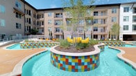 This expansive pool and lounge area is designed as a resort-style retreat, offering a well-appointed outdoor experience. The centerpiece is a large, freeform swimming pool with striking multicolored mosaic tile accents lining its edges, adding a bold and vibrant visual element to the space. A distinctive landscaped island rises from within the pool area, featuring native plantings, trees, and casual seating, creating a unique focal point.

A substantial covered pergola structure with slatted wood detailing provides a shaded lounge zone directly adjacent to the pool. This sheltered area is furnished with plush sectional sofas, accent chairs, patterned rugs, and side tables, making it well-suited for relaxed socializing. Ceiling fans enhance comfort under the pergola. An additional covered cabana features an outdoor television and bistro-style seating.

The surrounding pool deck is lined with rows of sun loungers and casual seating areas, offering plentiful space for sunbathing and outdoor recreation. A dedicated covered lounge area along the perimeter, enclosed by stone columns, provides a transitional indoor-outdoor space with additional soft seating. Accessible pathways and open walkways connect the various zones of the courtyard, which is framed by the surrounding multi-story building, cypress trees, and open sky.