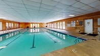 Located at 2005 Treehills Pkwy in Stone Mountain, Georgia, this single-story indoor pool facility offers a well-maintained aquatic space designed for lap swimming and recreational use. The centerpiece of the building is a full-length rectangular pool with clearly marked lap lanes, bordered by a generously sized concrete pool deck that provides ample space for entry, exit, and poolside activity.

The interior architecture is characterized by warm wood-paneled walls that run throughout the facility, lending a rustic yet welcoming aesthetic. A drop-tile ceiling with integrated lighting panels ensures consistent illumination across the pool area. Rows of large windows line the walls on multiple sides, allowing significant natural light to filter in while offering views of the surrounding wooded landscape — a notable feature that enhances the overall ambiance of the space.

An adjacent utility and entry area connects to the main pool room, featuring the same wood-panel detailing, additional doorways, and a large window providing outdoor views of mature trees. This transitional space also houses storage and maintenance equipment for pool upkeep. Double entry doors provide convenient access to the exterior. The facility is equipped with standard safety signage, a life ring, and poolside cleaning tools, reflecting its functional, community-oriented purpose.
