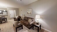 This single-floor space offers a warm and inviting atmosphere designed for comfortable everyday living. The living room serves as the heart of the space, featuring neutral beige and taupe tones throughout that create a calm, cohesive aesthetic. Plush wall-to-wall carpeting underfoot adds a sense of warmth and comfort to the room.

The seating area is anchored by a generously sized brown upholstered sofa accented with decorative throw pillows, complemented by a matching armchair that creates an ideal setting for relaxing or entertaining. A dark wood coffee table and coordinating side table provide practical surfaces, while two table lamps cast a soft, ambient glow throughout the room.

Framed artwork adorns the walls, adding a refined touch to the interior. The smooth, neutral-painted walls are accented by clean white baseboards and trimwork, contributing to the room's polished appearance. A recessed ceiling track light fixture provides additional overhead illumination.

The layout flows naturally, with a doorway visible in the background offering access to adjacent areas of the space. The overall design strikes a balance between functionality and comfort, making it a practical and welcoming environment suited for a variety of lifestyles.