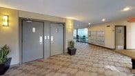 This building lobby offers a well-designed, functional entry experience characterized by a distinctive curved wall of large, floor-to-ceiling windows that flood the space with natural light and provide expansive views of the surrounding landscaped greenery. The generous open layout is finished with patterned wall-to-wall carpeting in neutral tones, contributing to a clean and inviting atmosphere.

A comfortable seating area near the curved window wall features dark leather accent chairs and a small side table, complemented by tall potted tropical plants that add a touch of warmth and life to the space. Recessed ceiling lighting supplements the abundant daylight, while the textured ceiling adds subtle architectural character.

Dual elevator doors serve as a central functional feature of the lobby, providing access to the building's floors. Adjacent to the elevators, a neatly organized bank of resident mailboxes lines the wall, offering convenient mail retrieval. A dedicated outgoing mail station is also present for resident use.

The lobby connects to an exterior courtyard area through sliding glass doors, creating a seamless transition between the interior and outdoor environments. Directional and unit-number signage is clearly posted, aiding navigation throughout the building. Overall, the lobby balances practicality with a light, open ambiance suited to a well-maintained residential building.