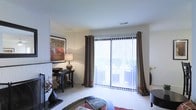 This single-floor space centers around a well-appointed living area designed for both comfort and functionality. The room's most prominent feature is a large sliding glass door framed by floor-length dark grommet curtains paired with sheer vertical blinds, allowing for flexible light control while providing access to an outdoor area. The generous glazing fills the space with natural light and offers views of surrounding greenery, enhancing the overall sense of openness.

The living area is finished with plush wall-to-wall carpeting in a neutral tone, contributing to a warm and inviting atmosphere. White walls serve as a clean backdrop, complemented by tasteful décor including a framed artwork piece and a classic table lamp atop a dark wood accent table. A patterned area rug anchors the seating zone, adding visual interest and texture to the space.

The layout is thoughtfully arranged to maximize usable floor space, with clearly defined areas for relaxing and entertaining. Recessed HVAC vents in the ceiling indicate a central climate control system. Overall, the space presents a clean, comfortable, and well-lit environment suitable for everyday living.