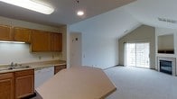 This single-floor residence showcases an open-concept design that seamlessly connects the kitchen and living area, creating a cohesive and functional layout. The kitchen is equipped with warm honey-toned oak cabinetry, a white dishwasher, and light-colored laminate countertops that extend into a peninsula or breakfast bar, offering additional prep space and casual seating. Recessed and flush-mounted ceiling lights provide ample illumination throughout the kitchen zone.

The living area transitions naturally from the kitchen, featuring neutral wall-to-wall carpeting that lends a comfortable, quiet atmosphere. A standout architectural element is the vaulted ceiling, which adds a sense of vertical spaciousness and openness to the room. Natural light filters into the living space through a sliding glass door fitted with sheer curtains, offering a view to the outdoors and providing a bright, airy feel. The overall color palette—white walls, light carpet, and warm wood tones—creates a clean, welcoming environment throughout the space.