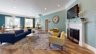 This single-floor space is designed as an open, multi-functional communal area with a cohesive aesthetic defined by soft teal walls, crisp white crown molding, and warm hardwood flooring that flows throughout. Recessed lighting overhead provides consistent illumination, complementing the natural light that enters through double glass doors and windows dressed with yellow curtains.

The main lounge area serves as the focal point of the space, anchored by a classic white fireplace with decorative ironwork. Comfortable seating — including a bold blue sofa and vibrant yellow accent chairs — is arranged around a patterned area rug, creating an inviting gathering environment. A wall-mounted television above the fireplace adds a functional element to the relaxed atmosphere. A large decorative clock serves as a notable wall feature.

Adjacent to the lounge, a bar-height counter lined with rich burgundy metal stools offers an informal seating area beneath warm pendant lighting. Curated artwork adorns the walls throughout, adding character and visual interest.

A separate room visible from the lounge contains a grand piano, suggesting a dedicated space for musical or creative activities. The entry foyer, accessible through glass-paned double doors, connects seamlessly to the rest of the space, reinforcing an open and welcoming flow.