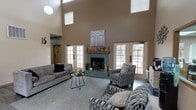 This single-story space is anchored by a spacious, open great room distinguished by dramatically soaring ceilings that rise well above typical height, creating an airy, grand atmosphere. Clerestory windows positioned high on the walls allow abundant natural light to filter in, complementing the warm, neutral taupe tones throughout. The focal point of the great room is a striking fireplace clad in teal-green tile, topped with a wooden mantel and decorative artwork. Multiple sets of French doors line the walls, providing views to the exterior and further enhancing the sense of openness.

The seating area features a mix of tufted sofas and patterned accent chairs arranged around a minimalist coffee table atop a large area rug, all set on durable wood-look flooring. Adjacent to the great room, a reception window with wood trim connects to a separate office area, indicating a professional or community-use function.

The kitchen is a practical galley-style space equipped with light oak cabinetry, a white gas range, refrigerator, and single-basin sink. A skylight overhead provides natural illumination. An exit door provides direct exterior access from the kitchen. Overall, the space balances a welcoming, comfortable lounge environment with functional support spaces suited for professional or community use.