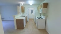 This single-floor residential space is thoughtfully laid out to maximize functionality and comfort. The kitchen serves as a central feature of the home, designed in a classic galley style that efficiently utilizes the available space. It is equipped with a full suite of white appliances, including a coil-burner electric range, over-the-range microwave, and a full-size refrigerator. Warm-toned wood cabinetry lines both sides of the kitchen, providing ample storage above and below the countertops. The countertops are finished in a light, neutral tone, complementing the overall color palette of the space. A single-basin sink is positioned along one side of the kitchen, accompanied by under-cabinet storage and a dishwasher. Light wood-style flooring runs through the kitchen area, transitioning to carpeting in the adjacent living areas, adding a sense of warmth and distinction between spaces. The walls are painted in soft, neutral tones, contributing to a clean and welcoming ambiance throughout. A door at the far end of the kitchen provides additional access, enhancing the flow and connectivity of the layout. Overhead flush-mount lighting ensures the kitchen remains bright and well-illuminated.