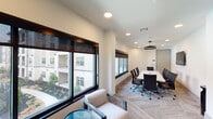 Located at 3200 Peek Rd in Katy, TX, this single-floor professional meeting space offers a sleek, contemporary environment designed to support productive collaboration. The layout is anchored by a spacious boardroom accommodating a large white rectangular conference table surrounded by ergonomic black woven-back chairs. A wall-mounted flat-screen display and a striking circular pendant light fixture serve as functional focal points, while recessed lighting throughout ensures consistent, professional illumination. Large dark-framed windows line one wall of the boardroom, inviting abundant natural light and offering views of the surrounding exterior landscape.

Adjacent to the boardroom is a comfortable lounge area featuring cream upholstered armchairs, a round blue glass accent table, and a decorative mosaic tile feature wall rendered in neutral tones of beige and grey — adding visual texture and a refined sense of style. The patterned carpet flooring in a herringbone-inspired layout flows consistently across both areas, contributing to the space's cohesive aesthetic.

Entry to the boardroom is through a set of sleek dark-framed glass doors, providing both visual transparency and a polished transition between zones. The overall design balances functionality with modern sophistication, making this space well-suited for meetings, presentations, and professional engagements.