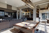 This thoughtfully designed single-floor amenity space blends contemporary style with inviting social functionality. Floor-to-ceiling windows line the perimeter, flooding the interior with abundant natural light and providing open views to an adjacent outdoor terrace. Large-format tile flooring anchors the main gathering areas, transitioning seamlessly between zones.

The central lounge area features a full-sized billiards table beneath a striking industrial-style pendant light fixture, accompanied by plush sectional seating and a wall-mounted large-screen television. Decorative striped wall treatments and curated artwork contribute to the space's upscale aesthetic. A recessed nook with a wraparound sectional sofa provides an intimate, semi-private retreat with layered wall textures and a tray ceiling detail.

A dedicated wet bar area includes dark cabinetry, a black countertop, under-counter beverage refrigeration, and a sink — ideal for entertaining. A sliding barn door adds an architectural accent nearby. The kitchen counter bar area features dark wood cabinetry with seating for multiple guests beneath pendant lighting.

Throughout, the design maintains a cohesive palette of warm browns, grays, and dark wood tones, creating a sophisticated and welcoming communal environment.