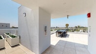 Located at 2600 S Sepulveda Blvd in Los Angeles, this rooftop terrace offers a thoughtfully designed outdoor amenity space with sweeping panoramic views of the Los Angeles skyline, surrounding neighborhoods, and iconic palm trees stretching to the horizon. The terrace is finished throughout with large-format, light-toned porcelain tiles that lend a clean, modern aesthetic and reflect the abundant Southern California sunshine.

The space is organized into several distinct functional zones. A dedicated sunbathing area features multiple chaise lounges arranged on green turf mats, providing a comfortable retreat for relaxation. A covered dining section offers a dark wood table with seating for several guests, partially shaded by an overhead structure and equipped with wall-mounted lighting for evening use. A fire pit lounge area presents modular outdoor seating arranged around a central table, ideal for social gatherings. An additional sectional sofa seating area offers generous, cushioned seating backed by the building's crisp white parapet walls.

The perimeter of the terrace is enclosed by white metal railings, ensuring safety while preserving unobstructed sightlines across the Los Angeles basin. Planters with greenery add natural accents throughout. The open-air design and elevated position make this rooftop an exceptional amenity, bathed in natural light and open to clear blue skies year-round.