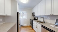 Located at 91 Front Ave W in Brockville, Ontario, this one-bedroom, one-bathroom unit offers a thoughtfully arranged single-floor layout. Warm-toned hardwood flooring runs throughout the living areas and bedroom, creating a cohesive and inviting atmosphere.

The kitchen is a standout feature, finished with crisp white cabinetry extending to the ceiling for ample storage, grey tile backsplash, and stone-look countertops. A full suite of stainless steel appliances — including a refrigerator, electric range, over-the-range microwave, and dishwasher — rounds out this functional cooking space. A window above the sink area allows natural light to fill the room.

The living room is open and airy, featuring a wide picture window that frames neighbourhood views and allows generous daylight to enter. Baseboard heating runs along the perimeter, providing efficient warmth throughout.

The bedroom is a comfortable, well-proportioned space with its own window offering outdoor views and continued baseboard heating. Clean white walls and hardwood flooring maintain the unit's consistent, neutral aesthetic.

The bathroom features floor-to-ceiling beige tile surrounding a soaker tub, complemented by a frosted privacy window and a white vanity with stone-look countertop. Overall, this unit presents a clean, modern, and functional living space.