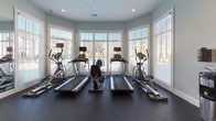 Located at 501 Hinson Dr in Myrtle Beach, South Carolina, this dedicated fitness center offers a well-equipped, single-floor workout facility designed to support a comprehensive exercise routine. The space is bathed in natural light, courtesy of large windows and glass doors that line one wall, offering pleasant views of the surrounding outdoor area and creating an open, airy atmosphere. The interior is finished with soft sage-green walls, clean white trim, and durable dark rubber flooring throughout, lending the space a modern yet functional aesthetic. A ceiling fan provides comfortable air circulation during workouts. The cardio zone features multiple treadmills and elliptical machines, all positioned to take advantage of the natural light and outdoor views. The strength training area is equally well-appointed, housing a multi-station cable and weight machine, a weight bench, and a full rack of dumbbells reflected in a large wall mirror that visually expands the space. Additional equipment includes medicine balls on a dedicated rack and a wall-mounted television for entertainment. A water dispenser and waste receptacle are conveniently placed near the entrance, and framed fitness-themed artwork adds a motivational touch to the walls. Recessed lighting supplements the natural light for early morning or evening use.
