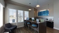 This business center is thoughtfully designed to provide a productive and comfortable environment for work. The space features a long, dark wood-toned counter workstation equipped with computer terminals, offering residents or visitors a dedicated area for professional tasks. Sleek bar-height seating with teal upholstered stools lines the counter, providing ergonomic and stylish workspaces. Overhead, a series of modern pendant lights illuminate the area with warm, focused lighting, complementing the overall contemporary aesthetic. Large windows with patterned fabric valances and horizontal blinds line one wall, allowing generous natural light to fill the room while framing views of the surrounding outdoor environment. Dark hardwood-style flooring anchors the space and adds a warm, polished finish. A vibrant blue abstract artwork mounted on a textured accent wall introduces a splash of color, contributing to the creative yet professional ambiance. The neutral, earth-toned walls and carefully curated décor, including small greenery accents on the counter, create an inviting atmosphere that balances functionality with modern design sensibility.