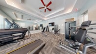 Located at 2841 Enterprise Rd in Orange City, Florida, this fitness facility offers a comprehensive workout environment spread across two distinct gym spaces on a single floor. The first gym area features a tray ceiling with a ceiling fan, patterned carpet flooring, and full-length wall mirrors that visually expand the space. This room is outfitted with Matrix-brand cardio equipment including treadmills and an elliptical, along with a dumbbell rack, adjustable benches, cable machines, and a wall-mounted flat-screen TV. Natural light filters in through windows fitted with blinds, creating a bright and energizing atmosphere. The second gym space offers a different aesthetic, with light wood-look vinyl plank flooring, soft sage-green walls, and a ceiling fan overhead. This room is equipped with a multi-functional cable machine, a dumbbell and medicine ball rack, plyo boxes, a stability ball, and a BOSU trainer, making it well-suited for functional and strength training. A large wall mirror enhances the open feel of this area. Together, the two spaces provide a versatile and thoughtfully designed fitness environment suitable for a wide range of workout styles and training needs.