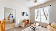 This well-designed 2-bedroom, 2-bathroom single-floor unit offers a thoughtful layout that makes efficient use of its space throughout. The combined kitchen and living area serves as the heart of the home, where the kitchen features warm honey-toned wood cabinetry lining both walls, white appliances including a refrigerator, range, microwave, and dishwasher, and a peninsula counter with bar seating that opens to the living room. The living room is bright and welcoming, with a ceiling fan overhead, wood-look plank flooring underfoot, and a large window dressed with sheer curtains that allows natural light to fill the room. A connecting hallway leads to the private quarters, where both bedrooms are carpeted and feature windows with blinds. The primary bedroom offers a comfortable footprint with ample natural light. Both bathrooms are finished consistently with warm wood vanity cabinetry, light countertops, wood-look flooring, and shower-tub combinations. A dedicated closet and additional hallway space with built-in hooks provide practical storage throughout. The neutral color palette of warm whites and beiges creates a cohesive and calm ambiance across the entire unit.