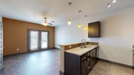 This single-floor unit offers a thoughtfully designed open-concept layout that seamlessly connects the kitchen and living area. The kitchen is a standout feature, equipped with dark espresso cabinetry, granite countertops, a peninsula with a built-in sink, and black appliances including a refrigerator, range, and microwave. Stylish pendant lighting in warm amber tones hangs above the peninsula, adding a distinctive visual accent to the space. Hardwood-style flooring runs throughout the main living areas, complementing the contemporary aesthetic.

The living area is bright and inviting, featuring a ceiling fan, a warm earth-toned accent wall, and a set of French doors that invite natural light and provide access to an outdoor area. A separate bedroom offers plush carpeting, a ceiling fan, and a broad window with expansive views of the surrounding area, allowing ample natural light to fill the room.

The bathroom is finished with tile-surround tub and shower combination, wood-style flooring, and granite countertops consistent with the rest of the unit's design. Throughout the space, textured walls and dark door and trim accents contribute to a cohesive and modern interior style. The overall ambiance is clean, functional, and contemporary.