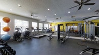 This fitness center offers a comprehensive workout environment designed to accommodate a wide range of exercise needs. The space features a generous open floor plan with durable rubber flooring, providing a safe and functional surface for all types of training. A row of cardio machines — including treadmills and an elliptical — lines one side of the room, while a dedicated strength training area with a cable machine and a full dumbbell rack occupies the opposite end. A bold yellow accent wall adds a vibrant, energizing touch to the space, complemented by large floor-to-ceiling mirrors that enhance the sense of openness and allow users to monitor their form. Natural light streams in through multiple windows along the walls, creating a bright and welcoming atmosphere. Ceiling fans are evenly distributed throughout, ensuring comfortable airflow during workouts. Recessed lighting provides consistent illumination throughout the day and evening. The overall ambiance is clean, modern, and motivating, making this fitness center a well-rounded facility suited for both cardio and strength-focused training routines.
