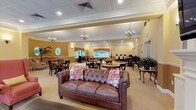 This single-story clubhouse offers a generously scaled, multi-zone gathering space designed to accommodate a variety of social activities. The interior is finished with warm golden-yellow walls, crown molding, and a combination of tile-patterned and carpeted flooring throughout, creating an inviting and refined atmosphere.

The main lounge area features a tufted leather sofa, coral wingback chairs, and a fireplace framed by a classic white mantel with a mounted television and built-in shelving above. Large windows dressed with plaid drapes allow abundant natural light and provide views of lush greenery outside. An adjacent rotunda-style sitting area is defined by a curved wall with oval windows, a chandelier, and elegant upholstered seating surrounding a pedestal table.

The dining and social areas include multiple round bistro-height and standard tables with dark wood chairs, ideal for casual dining or games. A dedicated space houses a full-size pool table. Tall-backed round-top double doors at the entrance are flanked by symmetrical plant displays, reinforcing the clubhouse's traditional charm. A kitchen is accessible nearby, supporting events and gatherings throughout this well-appointed community space.