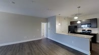 This single-floor residence features an open-concept design that seamlessly connects the living area to the kitchen. Dark hardwood flooring runs throughout the main living space, providing a rich contrast against the soft gray walls and bright white baseboards and trim. The layout feels spacious and unobstructed, allowing for flexible furniture arrangement within the living area.

The kitchen is positioned along one side of the open plan and is defined by a peninsula counter finished with granite-look surfaces, which also houses a sink. Dark cabinetry overhead adds depth, while pendant lights hang above the counter, offering both task lighting and a decorative element. A black refrigerator anchors the kitchen's appliance lineup. A door adjacent to the kitchen leads to an additional room or utility area, adding further functionality.

Smooth, neutral-toned ceilings with recessed lighting fixtures illuminate the space evenly. A doorway visible from the main living area provides access to other parts of the unit, suggesting a practical, well-organized floor plan. Overall, the space presents a clean, modern aesthetic well-suited for everyday living.