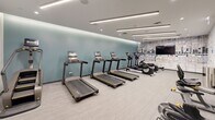 Located at 1717 Ridge Ave in Evanston, Illinois, this modern fitness center spans a single floor and is divided into two dedicated exercise rooms, offering a comprehensive and well-appointed workout environment. The space features a clean, contemporary design characterized by neutral grey tones, sleek linear LED lighting embedded in the ceilings, and light-toned carpet flooring throughout.

The cardio-focused room is anchored by a striking teal accent wall and houses a row of treadmills, elliptical trainers, and a stair climber, all positioned for ease of use. A wall-mounted flat-screen TV adds to the experience for those engaged in longer cardio sessions.

The strength and functional training room is equally well-equipped, featuring a power rack, a full dumbbell rack, cable machines, TRX suspension trainers, and padded workout mats. Full-length mirrors line one wall to support proper form during strength training. An additional area accommodates spin bikes, stability balls, a BOSU trainer, and other functional fitness accessories, rounding out the variety of training options available.

The overall ambiance is bright, clean, and professional, with recessed lighting and linear fixtures ensuring the space is well-illuminated. The thoughtful layout and diverse equipment selection make this fitness center suitable for a wide range of workout routines.