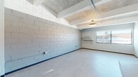 This single-floor space showcases an industrial-modern aesthetic defined by raw architectural elements alongside contemporary finishes. The main open area features prominently exposed concrete block walls painted in a light grey tone, polished concrete floors, and a ceiling with visible structural beams and acoustic tile panels. Overhead fluorescent and flush-mount lighting fixtures illuminate the generous open floor area, while a wall-mounted HVAC unit provides climate control. A window in this main zone allows natural light and exterior views to enter the space.

The kitchen area is equipped with a peninsula countertop featuring dark lower cabinetry contrasted by light wood upper cabinets, a stainless steel sink, microwave, and a black refrigerator. Soaring ceiling height in this section accentuates the open, loft-like character of the space.

Additional private rooms feature smooth grey drywall finishes, dark baseboards, polished concrete floors, and windows that invite natural daylight and outdoor views. The bathroom includes a dark wood vanity with an integrated sink, a toilet, and a shower-tub combination, with smooth grey walls throughout.

Overall, the space presents a clean, utilitarian industrial character with modern functional fixtures throughout.
