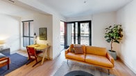 This thoughtfully designed single-floor residence offers a cohesive open-plan layout with consistent light hardwood flooring that flows seamlessly from room to room, creating a warm and airy atmosphere throughout.

The living area serves as the heart of the space, bathed in natural light from large black-framed glass doors that open to a private balcony with street views. The room comfortably accommodates a seating arrangement and includes a dedicated nook for a home office or study area, complete with a desk and ample wall space for personalization.

The kitchen is a standout feature, showcasing a two-tone cabinetry design with dark lower cabinets and white upper cabinets, accented by a textured mosaic tile backsplash. Brass-finished fixtures add a refined touch, while stainless steel appliances — including a refrigerator, electric range, and over-the-range microwave — provide full functionality. Open shelving offers additional display and storage options.

The bathroom is finished with classic hexagonal white floor tiles and full-height square wall tiles surrounding the bathtub and shower combination. A dark vanity with an undermount sink and a large mirror complete the space with a clean, modern aesthetic.

Overall, the unit presents a contemporary and well-appointed interior with a clean, minimalist design sensibility balanced by warm tones and curated details.