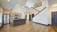Located at 2136 Central Ave in Indianapolis, IN, this thoughtfully designed two-story home offers three bedrooms, two bathrooms, and a generous, well-appointed layout. The main floor impresses immediately with soaring double-height ceilings in the open-concept kitchen and living area, where large windows flood the space with natural light. The kitchen features a central island with a built-in sink, stainless steel appliances including a range, microwave, and refrigerator, complemented by taupe cabinetry and subway tile backsplash. Warm hardwood-style flooring runs throughout the main level, adding continuity and a modern aesthetic. A dedicated laundry room with full-size washer and dryer adds everyday convenience. The primary bedroom is a spacious retreat with corner windows offering neighborhood views, a ceiling fan, and access to a well-finished en-suite bathroom complete with a dual-sink vanity, ample cabinetry, and a glass-enclosed shower. Additional bedrooms are bright and airy, each with large windows. The upper floor, accessed via a staircase with sleek dark wood railings, features plush carpeting and an open loft-style hallway overlooking the main level. Three closets throughout the home provide excellent storage.