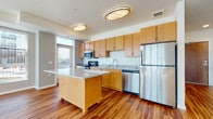 This single-floor residence offers a well-designed open-concept layout that seamlessly connects the kitchen and living areas. The kitchen is a standout feature, equipped with stainless steel appliances including a refrigerator, range, microwave, and dishwasher. Light maple cabinetry provides ample storage, while granite countertops extend across both the perimeter surfaces and a central island, offering generous workspace and a casual dining option. Rich hardwood flooring flows throughout the main living areas, adding warmth and continuity to the space.

The expansive living area is bathed in natural light, courtesy of a large bank of floor-to-ceiling windows dressed with vertical blinds, offering views of the surrounding urban landscape. The open layout creates a spacious, airy ambiance ideal for both everyday living and entertaining.

The bedroom features soft carpeting, large windows that invite abundant natural light, and a closet with access to additional storage. The bathroom is clean and functional, featuring a maple vanity with a white countertop, a large mirror, and a tiled tub-shower combination. Grab bars and thoughtful accessibility features are present throughout the bathroom, enhancing comfort and safety. Consistent hardwood flooring in the bathroom ties the unit together with a cohesive aesthetic.