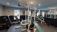 Located at 26 Harvard Rd in Ayer, MA, this single-floor fitness facility is designed to support a comprehensive workout experience. The space is divided into distinct functional zones, each purpose-built for different training modalities. The cardio area houses multiple upright stationary bikes and an elliptical trainer, all positioned along a two-tone wall featuring a dark lower wainscoting paired with a light upper section. Rubber flooring throughout the facility provides durability and safety underfoot. A dedicated strength training area centers around a substantial multi-station cable machine, complemented by a treadmill and a dumbbell rack stocked with a full range of free weights. Full-length mirrors line one wall of the strength zone, visually expanding the room and allowing users to monitor their form during exercise. Recessed ceiling lighting provides consistent, well-distributed illumination across the space. Multiple electrical outlets are positioned along the walls for added convenience. Natural light enters through several windows fitted with blinds. The overall ambiance is functional and utilitarian, with a clean, gym-appropriate aesthetic suited for focused training.