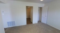 This single-story space presents a straightforward and functional layout designed to maximize practicality. The bedroom is finished with wall-to-wall carpet in a neutral beige tone, contributing to a warm and understated ambiance throughout. The walls are painted crisp white, creating a clean, bright atmosphere that feels open and easy to personalize.

A notable feature of this room is the dual closet configuration. A walk-in closet is accessible through an open doorway, offering generous storage space with visible shelving and hanging rods, making it well-suited for organizing clothing and accessories. Adjacent to it, a second open closet provides additional built-in shelving, further enhancing storage capacity for the room.

A wall-mounted ventilation register is positioned near the baseboard on one side of the room, indicating a centralized heating and cooling system. The ceiling features a flush-mounted light fixture, providing straightforward overhead illumination. Overall, the space presents a neutral, move-in-ready environment with an emphasis on clean lines, ample storage, and functional design.